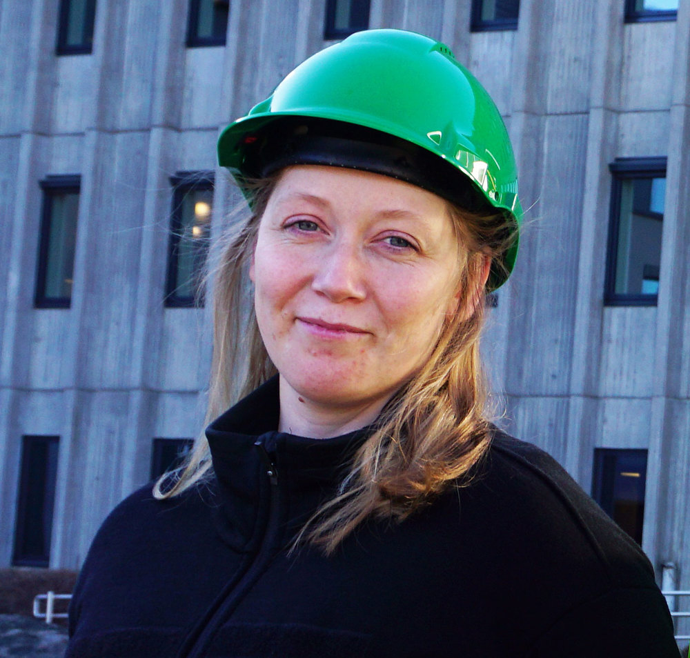 kvinne, langt hår, grønn hjelm og mørk jakke, betongbygg i bakgrunnen med masse vinduer.