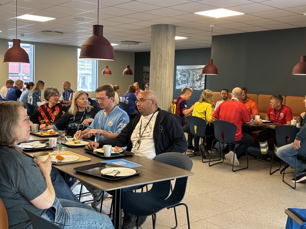 mennesker som sitter i kantine og spiser, mange har på fotballtrøye