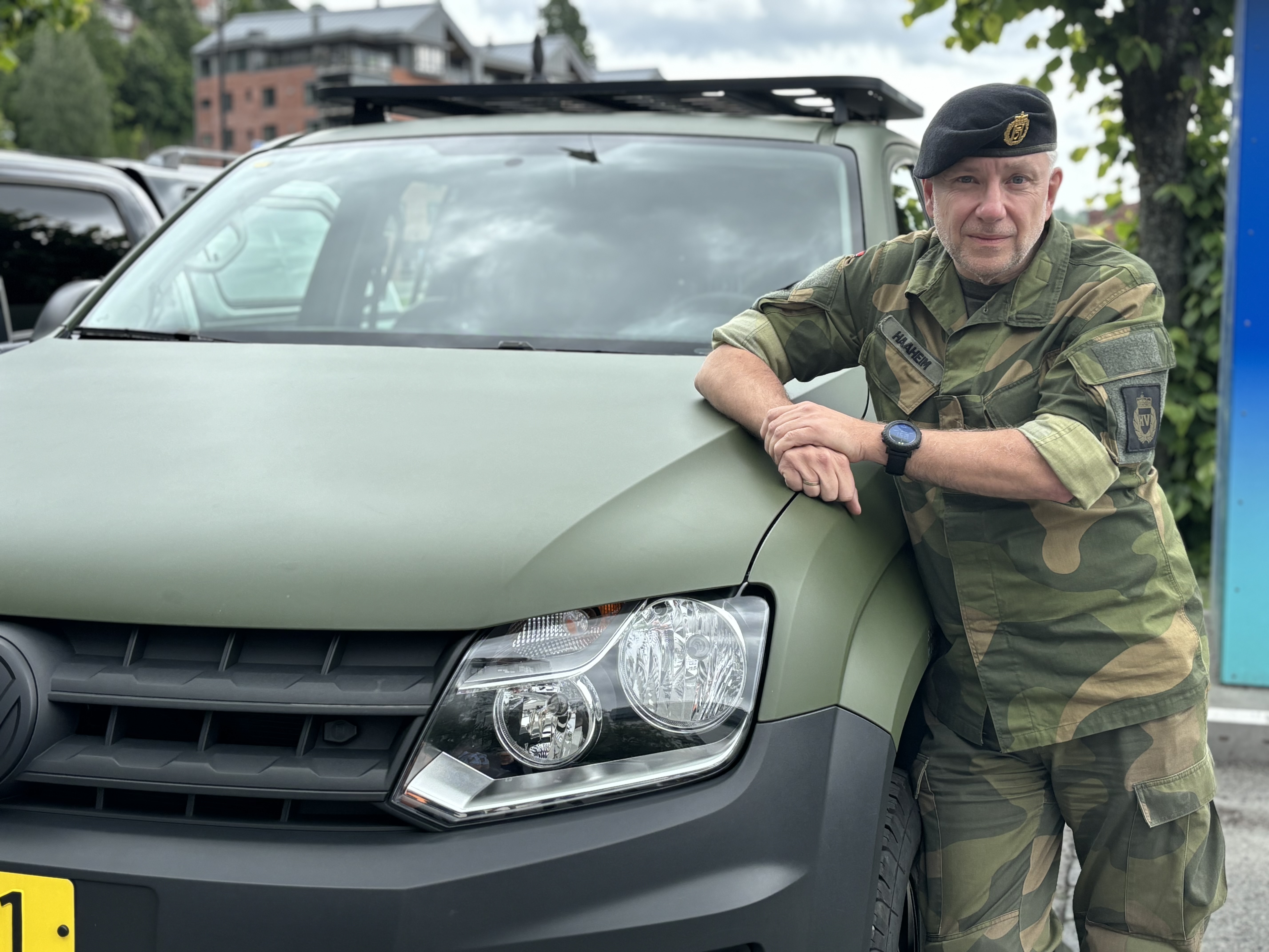 mann kledd i militær uniform lener seg mot et militært kjøretøy, poserer, bymiljø i bakgrunnen
