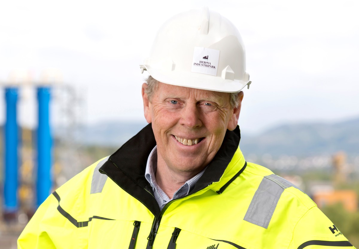 portrett av mann i hvit hjelm og gul jakke, smiler, industripark i bakgrunnen