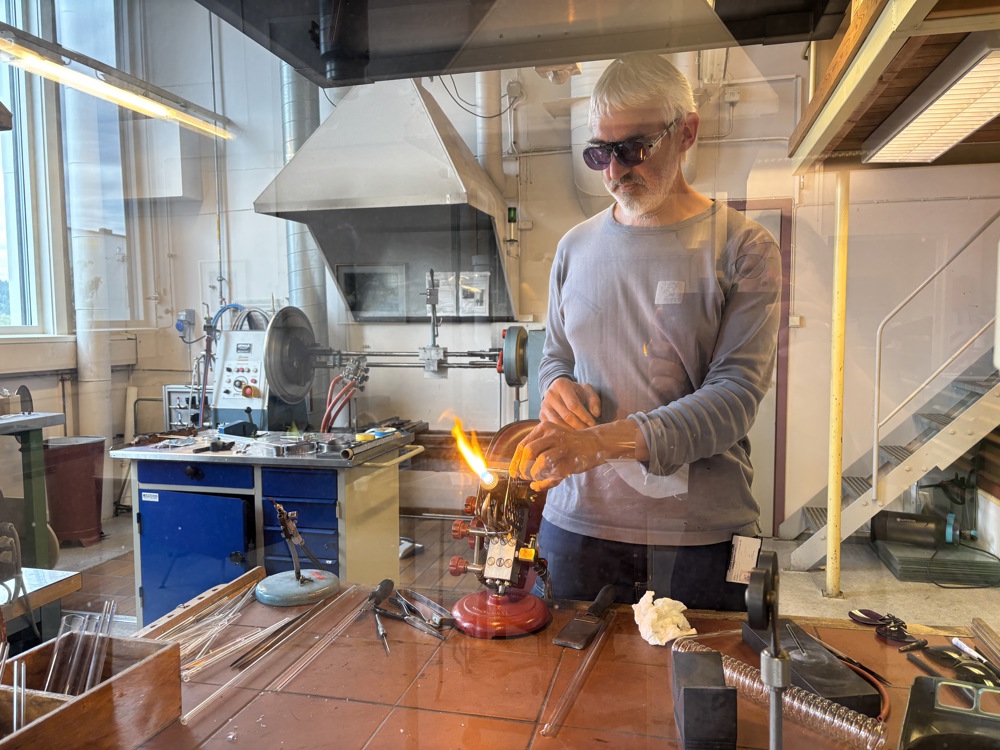 glassblåser i arbeid bak en glasskjerm, åpen flamme