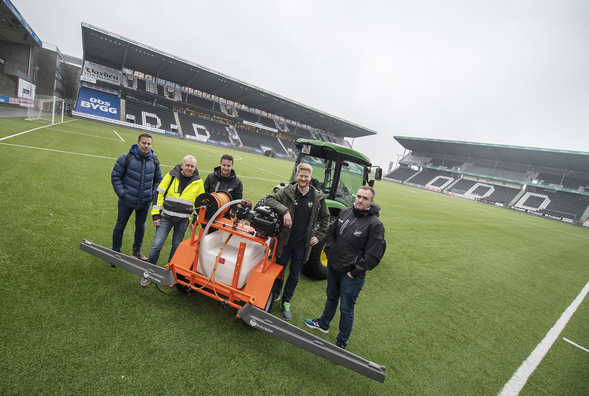 fem personer ute ved traktor på kunstgressbane for eliteseriefotball