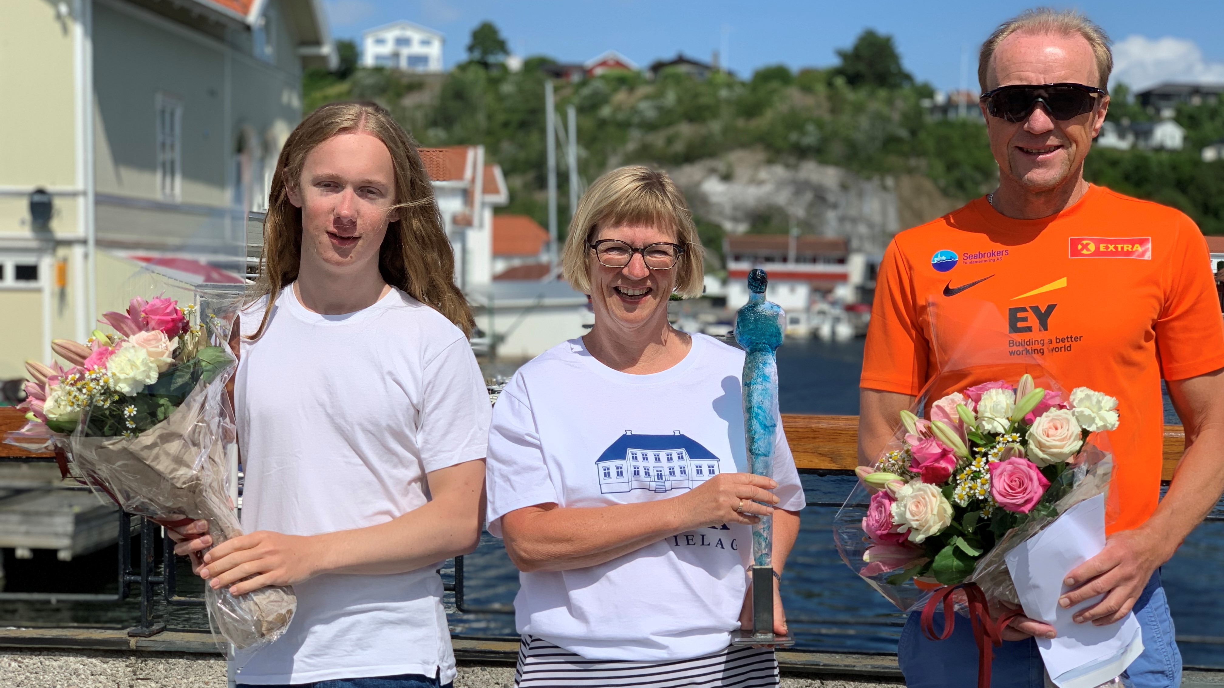 Tre personer står på en bro med sjø og bebyggelse i bakgrunnen. En ung mann til venstre holder blomster, en kvinne i midten holder en glasskunst og en høy mann til høyre holder blomster. 