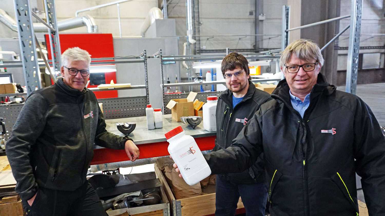 tre menn poserer, står i et verksted, en av dem holder en hvit plastbeholder med rødt lokk i hånden.