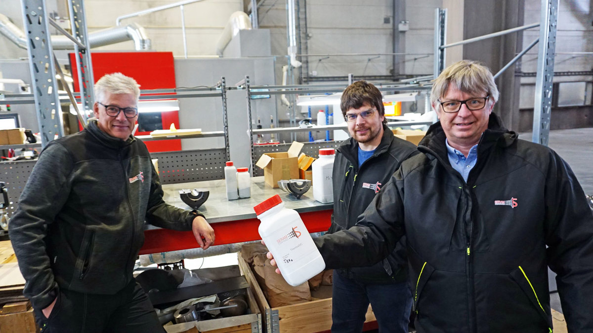 tre menn poserer, står i et verksted, en av dem holder en hvit plastbeholder med rødt lokk i hånden.