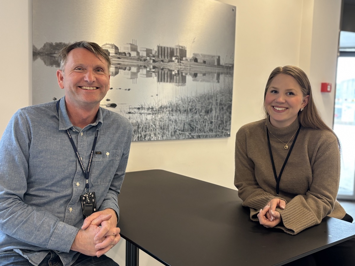 to personer lener seg inn over et bord. poserer, smiler. i bakgrunnen et trykk på aluminiumsplate.