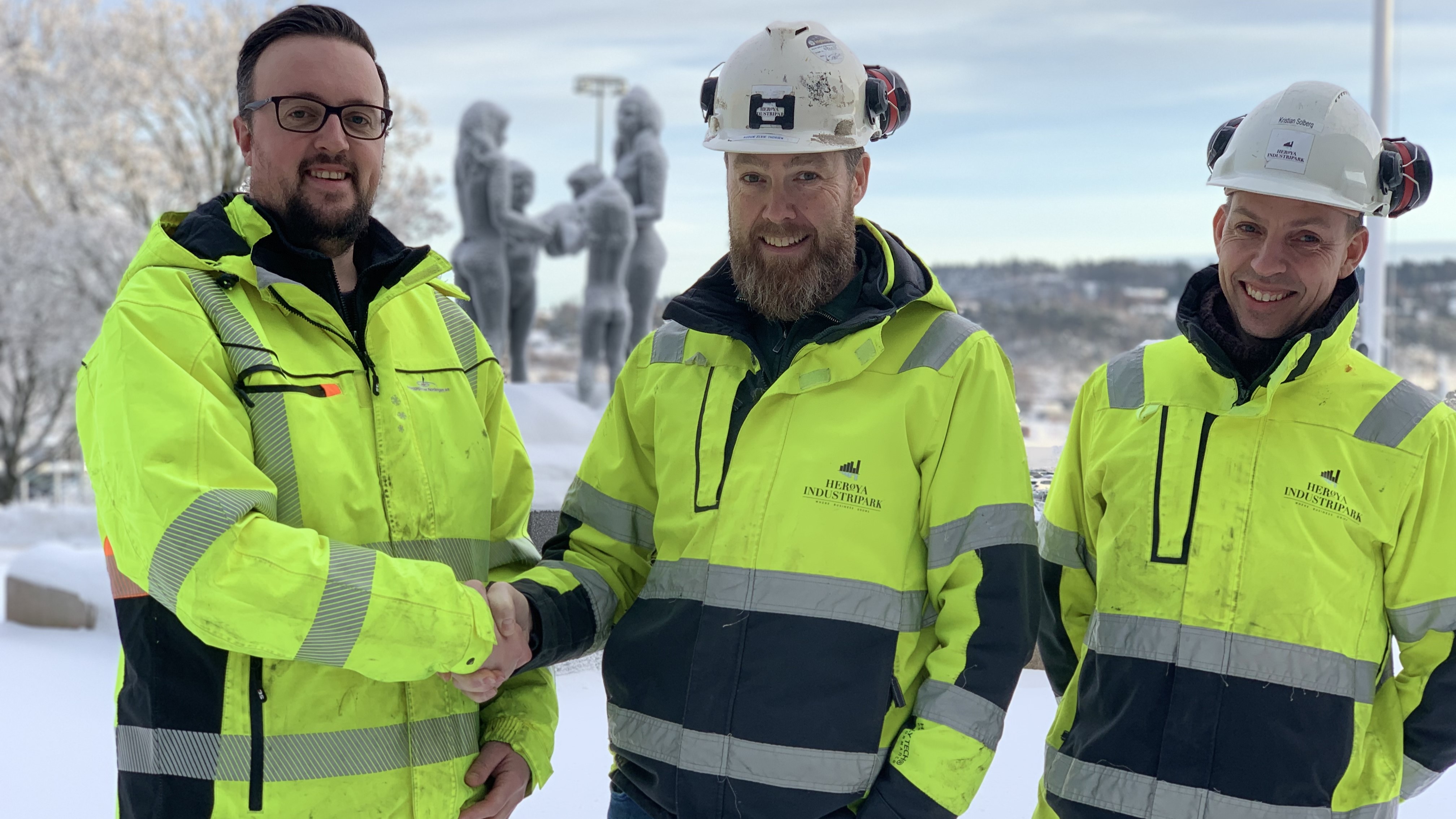 tre menn i gule jakker og hjelm på hode står ute i snø med bronsefigurer og parkanlegg i bakgrunnen