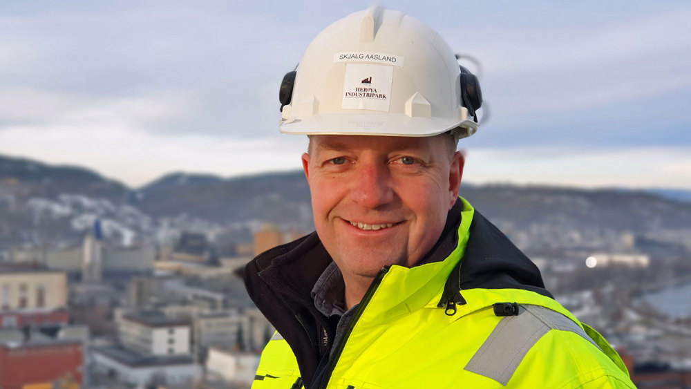 portrett av mann, smiler, poserer, hvit hjelm, står på en høyde, industripark i bakgrunnen