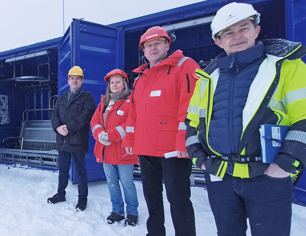 fire personer står ute i snø foran blå container