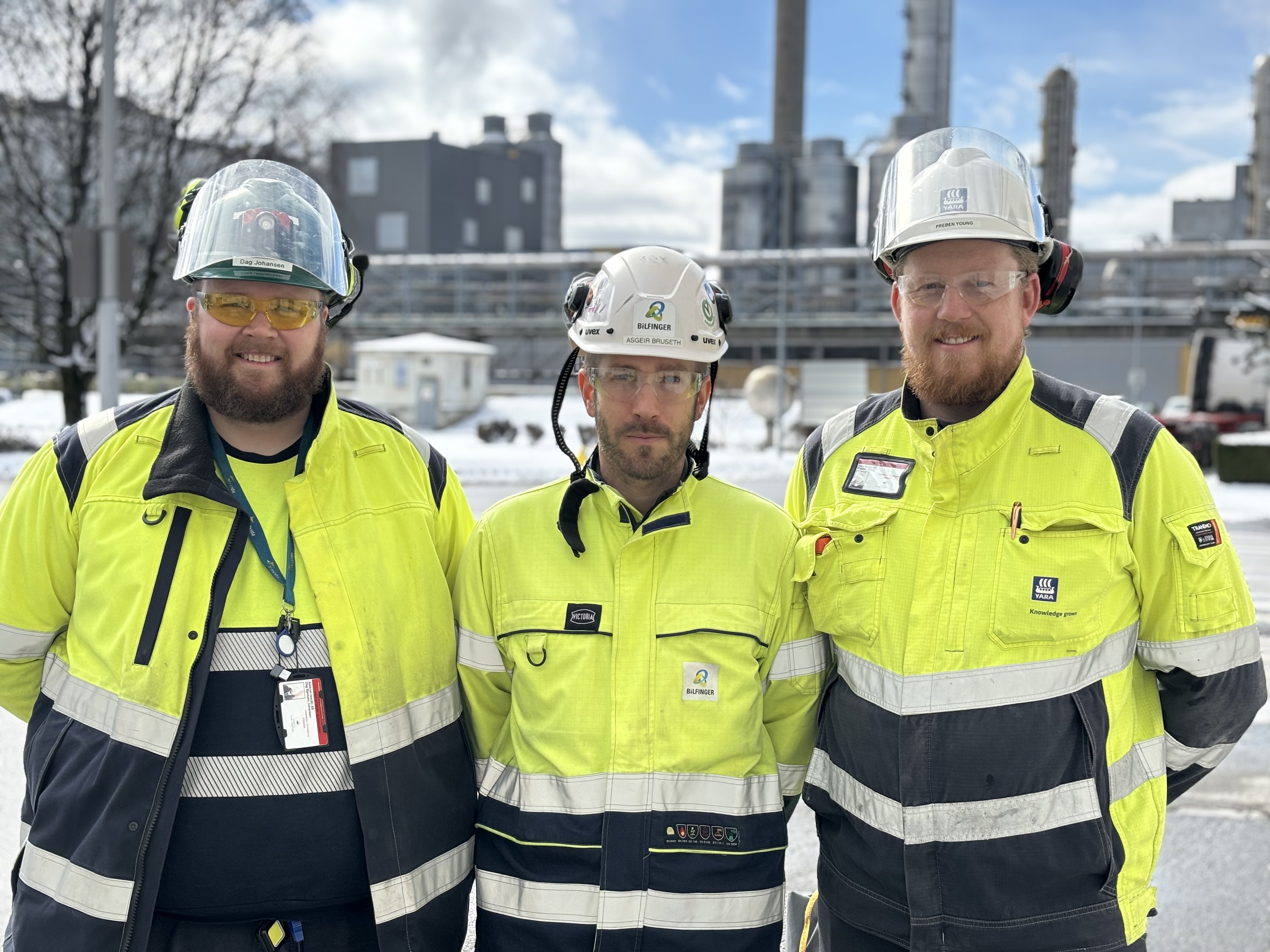 three persons, posing, standing close together, wearing PPE, back against the industrial park