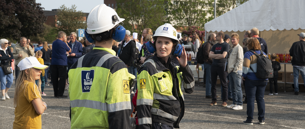 to Yara-ansatte med arbeidsklær og hjelm og øreklokker besøker festivalplassen