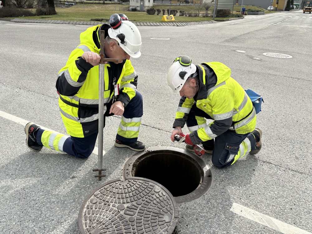 to menn sitter ved et åpent kumlokk i en gate. Begge kledd i verneutstyr. En mann holder kamera og tar bilde ned i kummen, den andre holder et spett som åpner kumlokk.