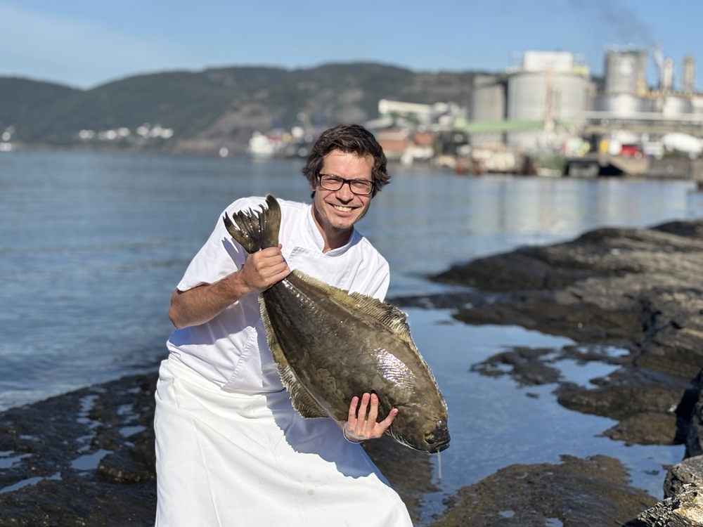 chef, posing, holding a big flat fish, industry and sea in background