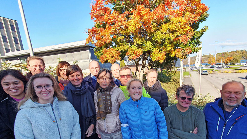 group of 12 people, smiling, posing, outside near a tree with autumn coloured leafes, and a car park behow.