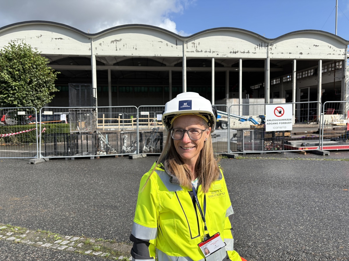 kvinne, poserer, i gul jakke og hjelm foran byggeplass, rehabilitering av stort murbygg