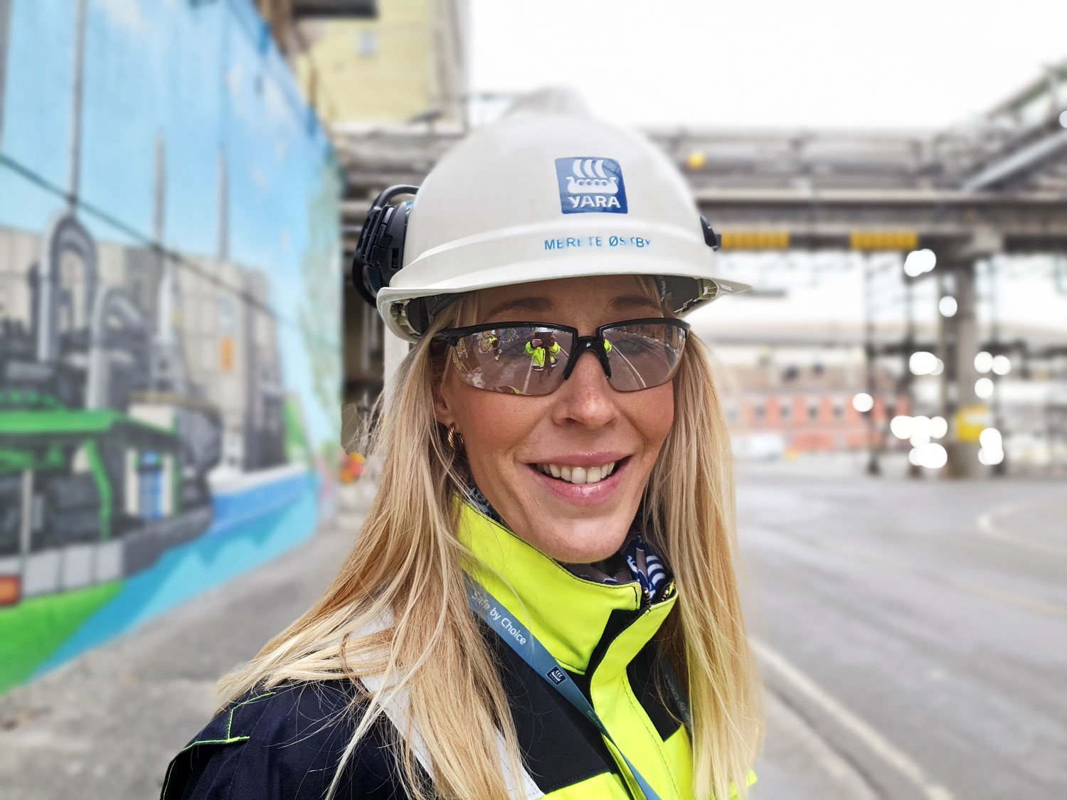 portrett av kvinne, langt blondt hår, hvit hjelm, blå og gul jakke, vernebriller, hun står i en gate i en industripark, dekorert betongvegg i bakgrunnen.