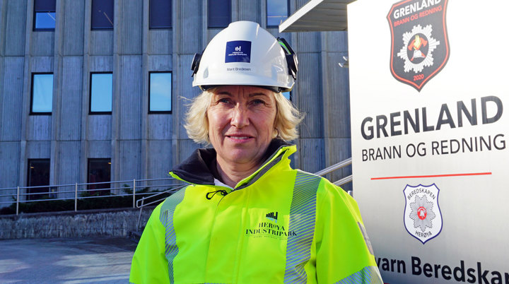 portrett, kvinne, poserer, gul jakke, hvit hjelm, firmaskilt bak, og stort kontorbygg i bakgrunnen