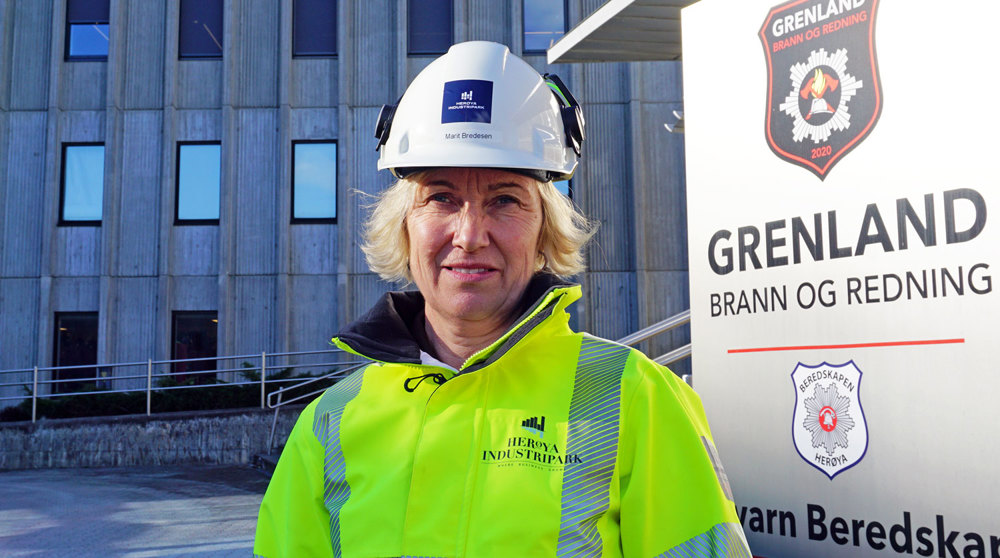 kvinne lyst hår, hvit hjelm, gul jakke, poserer ved siden av skilt med bedriftslogoer, ute foran betongbygg