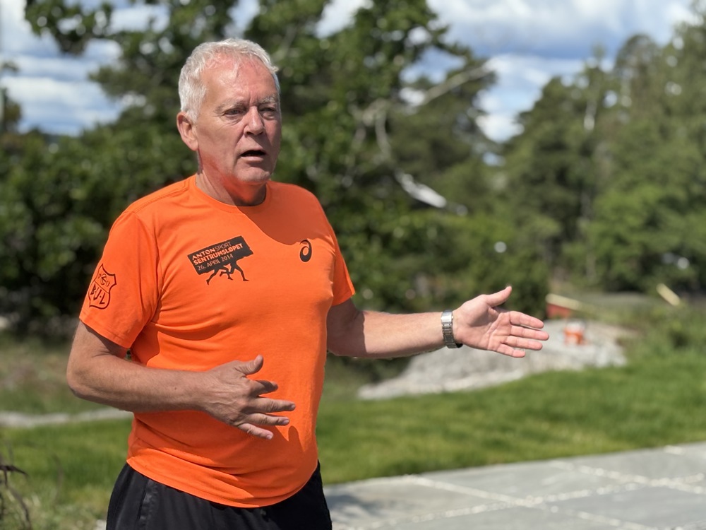 man in orange t-shirt, gesticulating, talking, green background and blue sky.