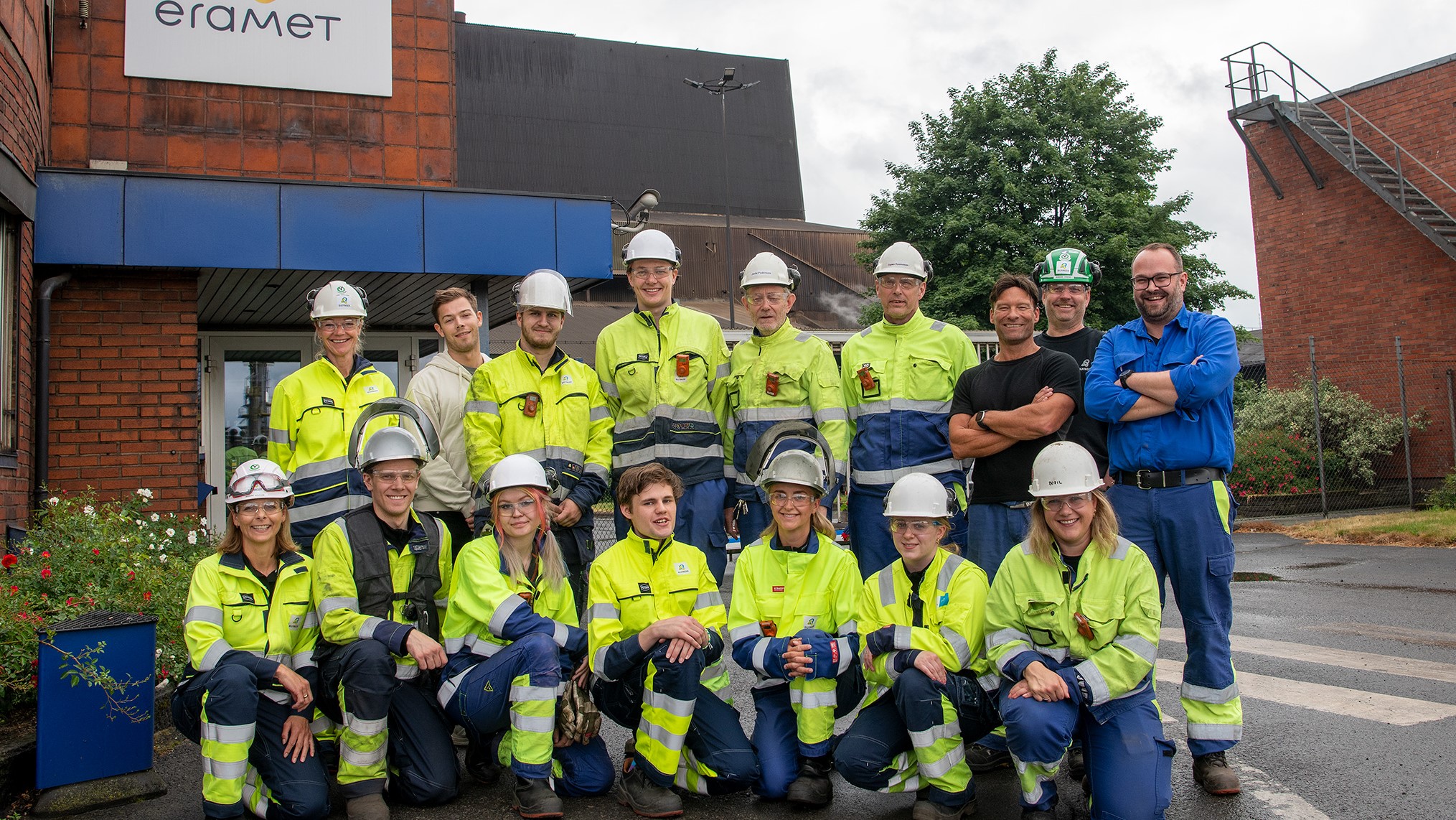 klassebilde. Stor gruppe med sommervikarer organisert som klassebilde, alle kledd i gule og blå arbeidsklær og hvite hjelmer. Utenfor inngangsport til fabrikkanlegg.