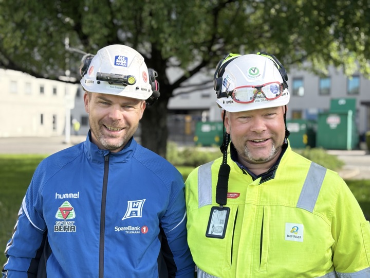 to glade menn med hjelm og gul og blå jakke, poserer, grønne trær i bakgrunnen