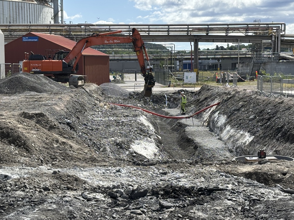 excavator in operation, large ditch, industial park, pipe bridges in background.