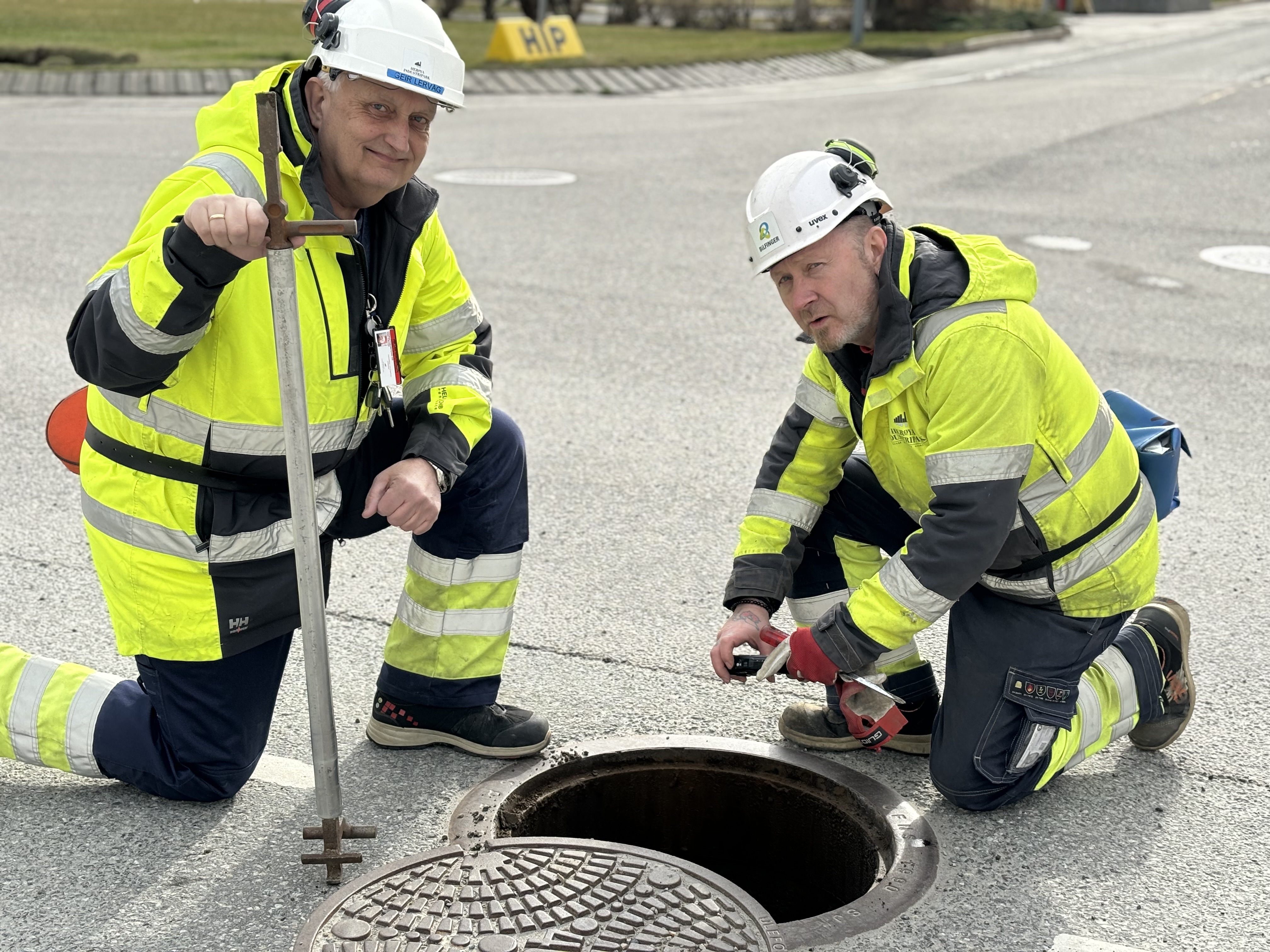 to menn sitter på huk ved en kum i gata, kumlokket ligger ved siden av. en mann holder et spett som kumlokket åpnes med, den andre bruker kamera ned i kummen og tar bilde.