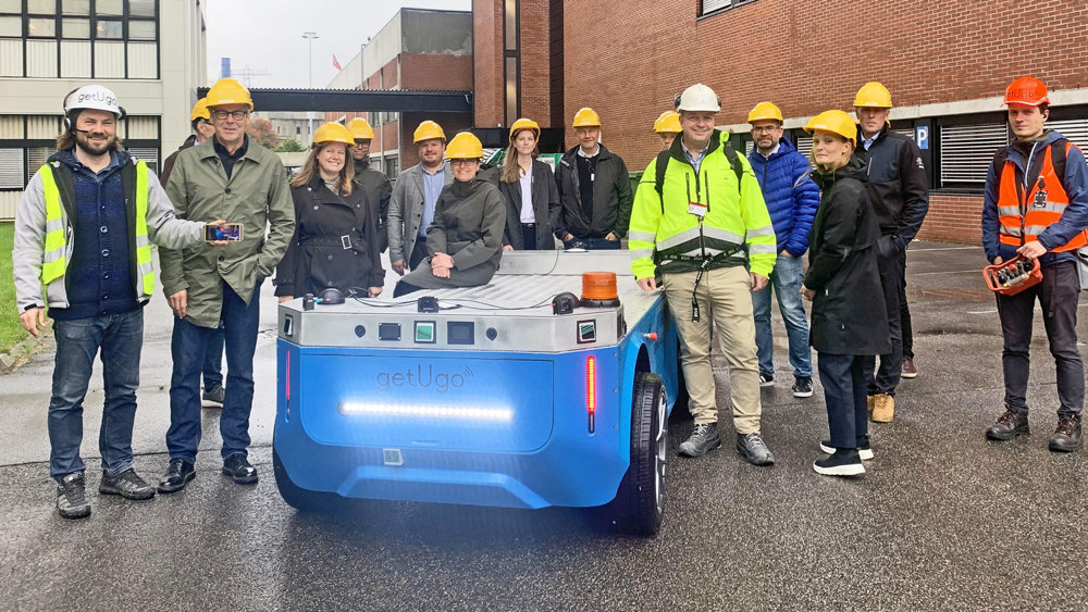 Approximately 15 people standing outside, posing, some of them leaning towards a one meter tall autonomous blue vehicle. Industrial park, they are all wearing yellow and white helmets.