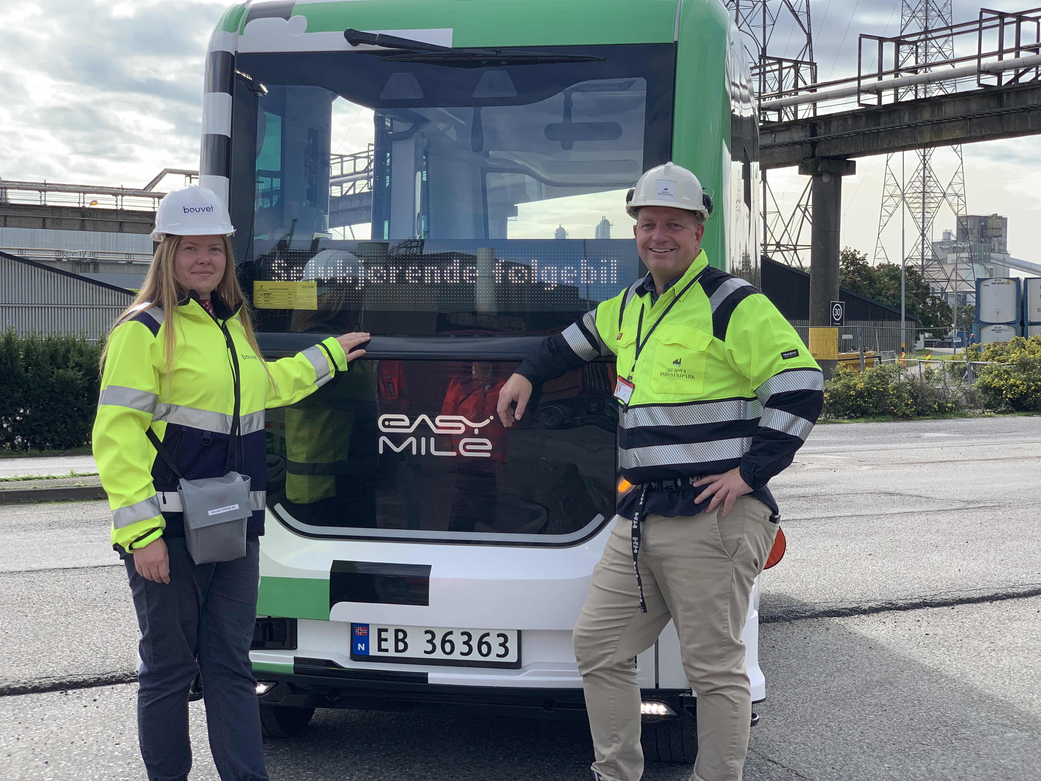 To personer poserer foran og lener seg mot en autonom minibuss, som er hvit og står parkert i industriparkomgivelser.