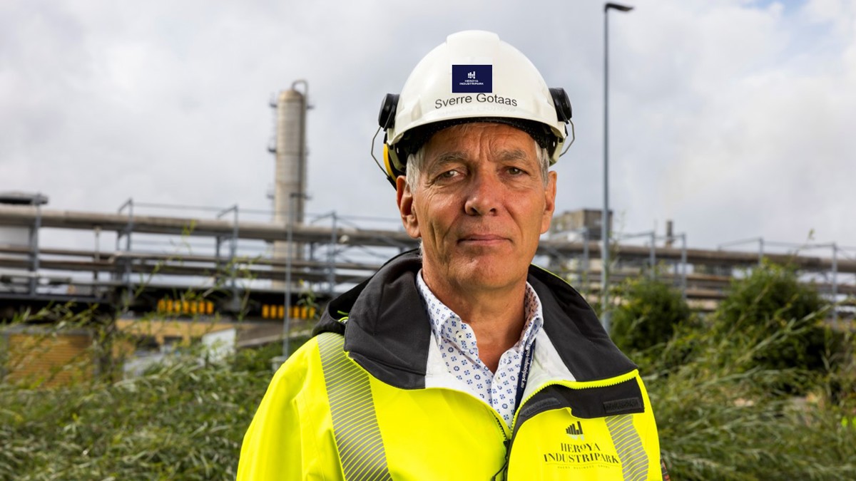 portrett av mann, hvit hjelm, gul jakke, fabrikkpiper i bakgrunnen