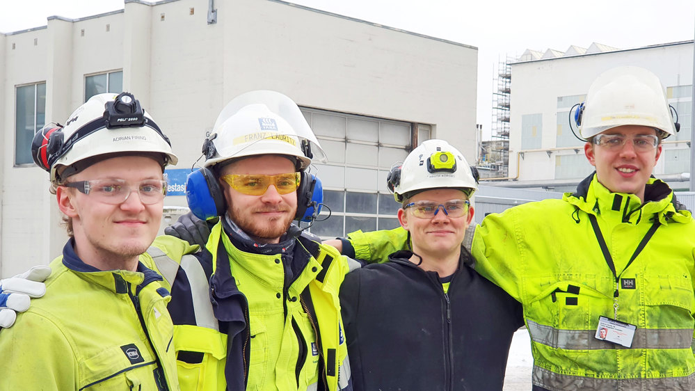 fire unge menn står på rekke, poserer med armene på hverandres skuldre, hvite hjelmer og verneutstyr.