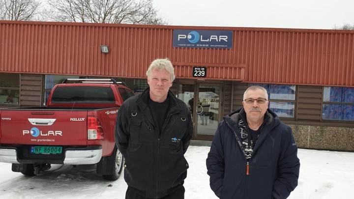 to menn står ved siden av hverandre foran et lavt verkstedbygg, med logo