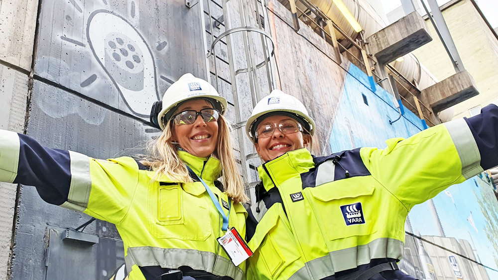 To unge kvinner poserer,  i arbeidsantrekk og hvite hjelmer, utstrakte armer, industrimiljø