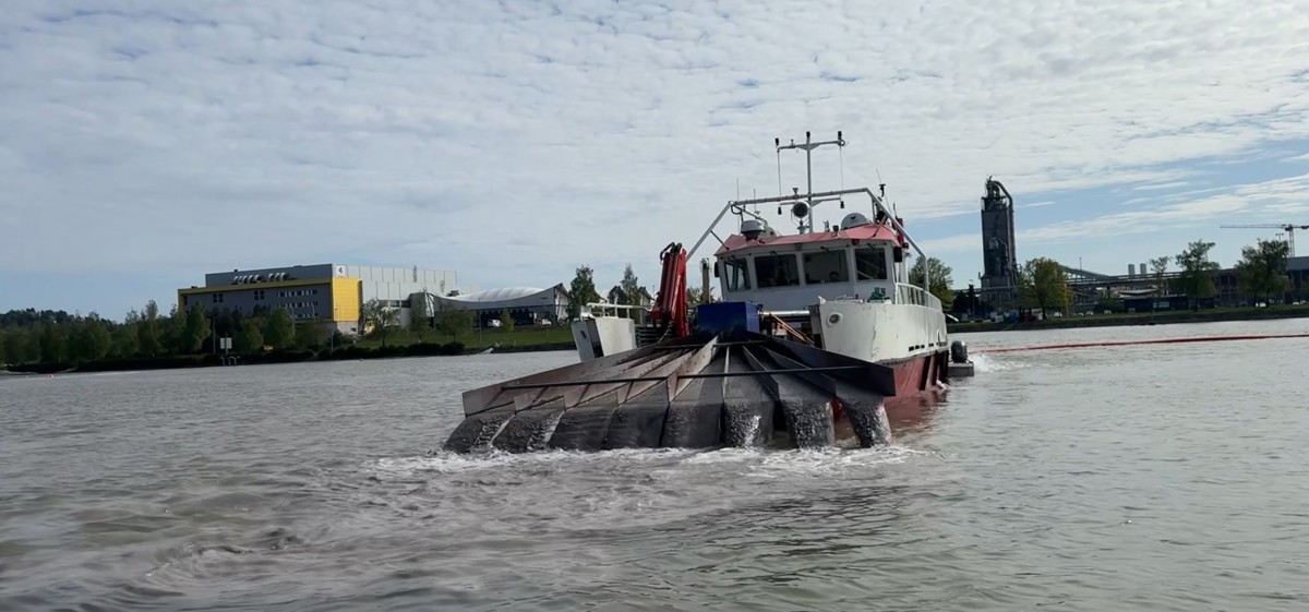 båt med utstyr i baugen som fordeler småstein og vann ut i innsjø. industripark i bakgrunnen.