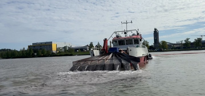 båt med utstyr i baugen som fordeler småstein og vann ut i innsjø. industripark i bakgrunnen.