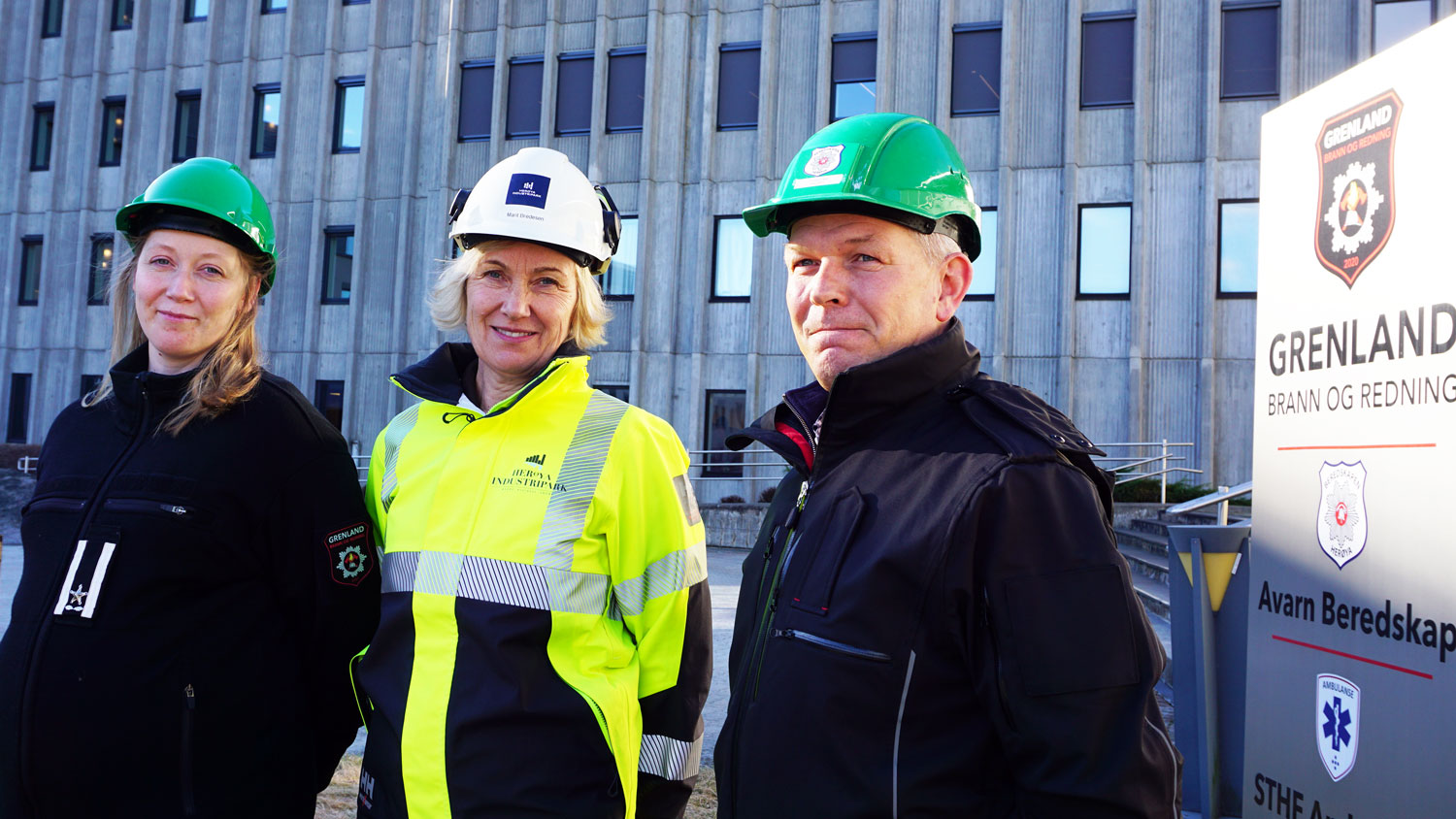 tre personer poserer, arbeidsjakker og hjelmer, betongbygning med masse vinduer i bakgrunnen og pylon med logoer på ved siden av personene