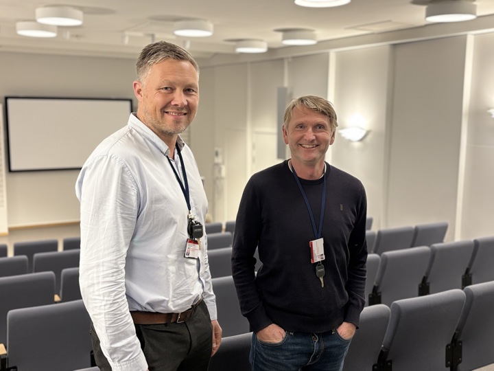 two men posing, auditorium, rows of chairs, screen on wall in background