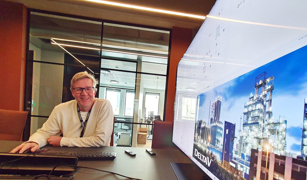 man, sitting, posing, desk, monitor on one wall, glass wall behind him.