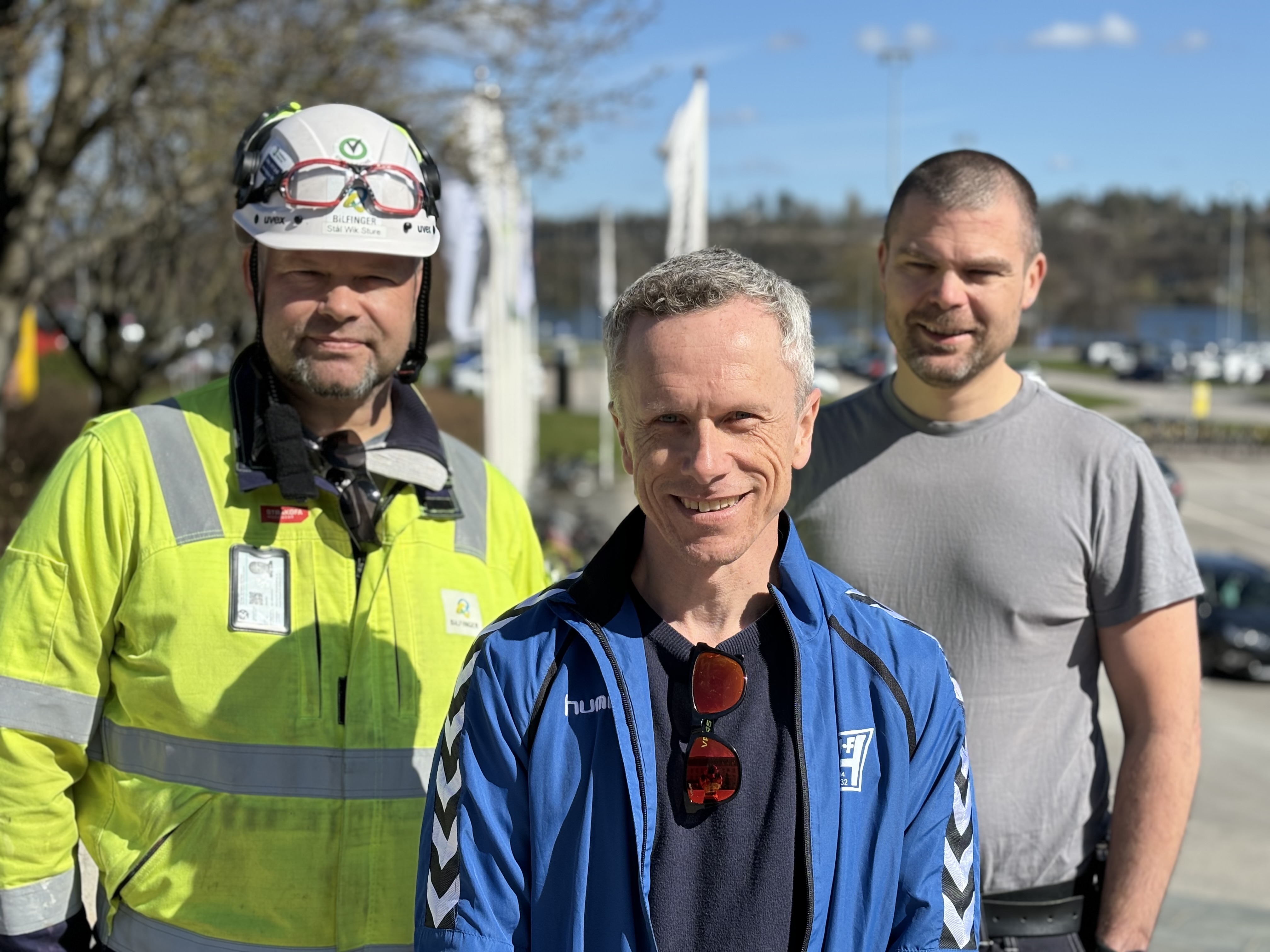 tre menn poserer, står tett sammen, en kledd i arbeidstøy, to kledd i sivil, parkeringsplass i bakgrunnen. Den ene mannen står litt foran de to andre.