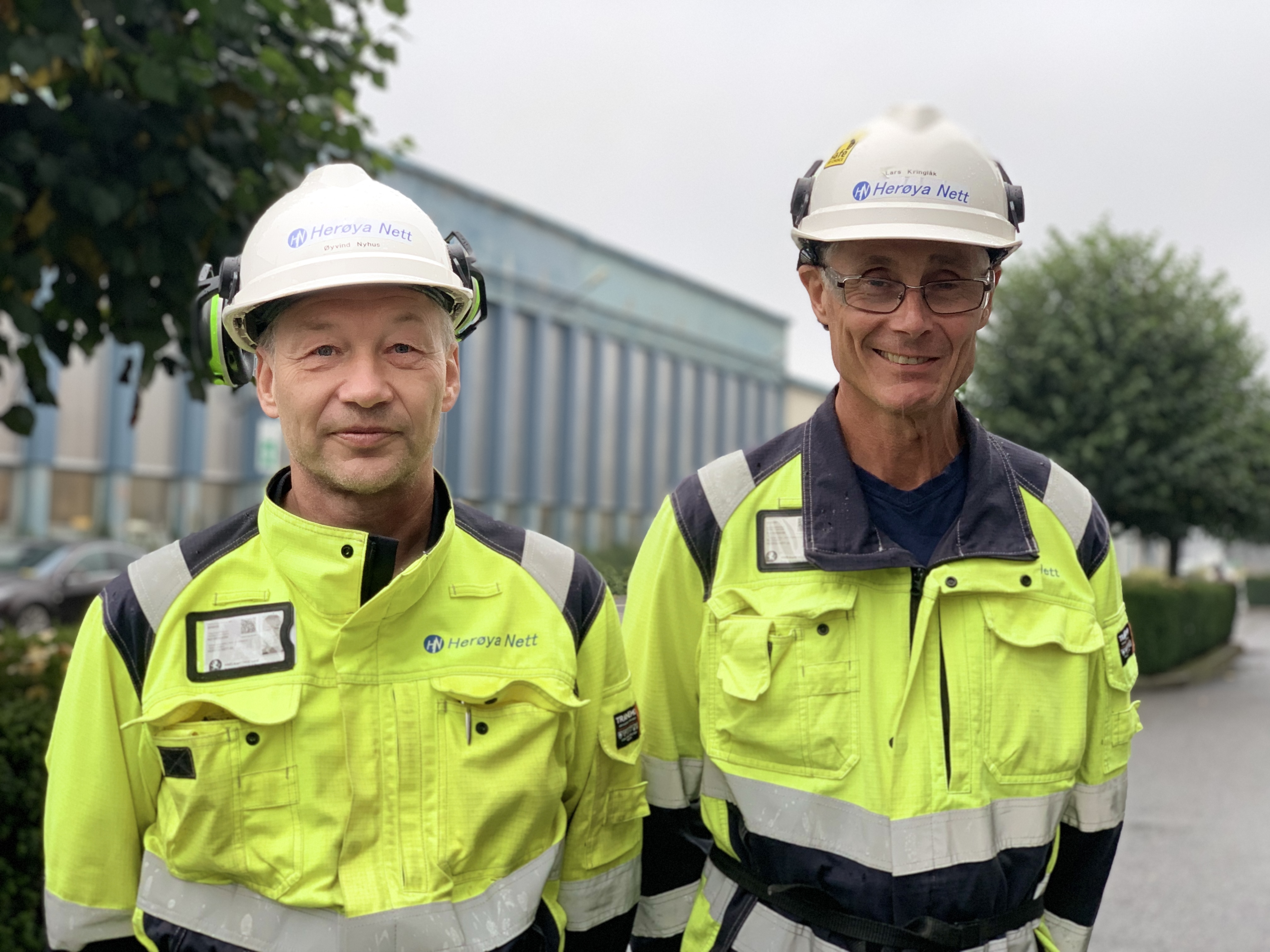 to menn står ved siden av hverandre, poserer, gule jakker, hvite hjelmer, i gate i industripark, grønne trær og industribygg i bakgrunnen.