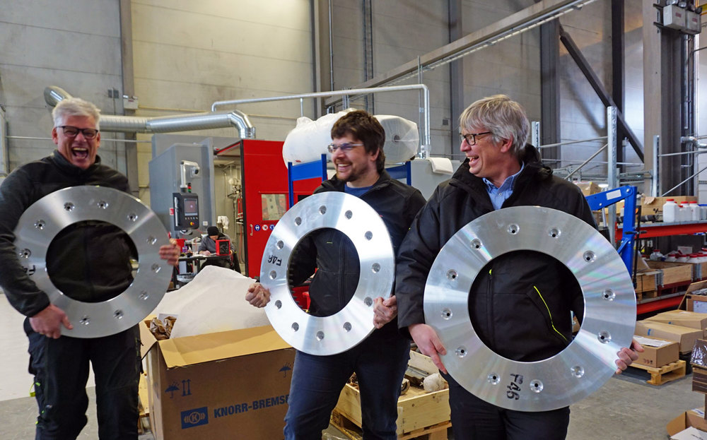 three men holding and displaying aluminium break discs for trains, workshop