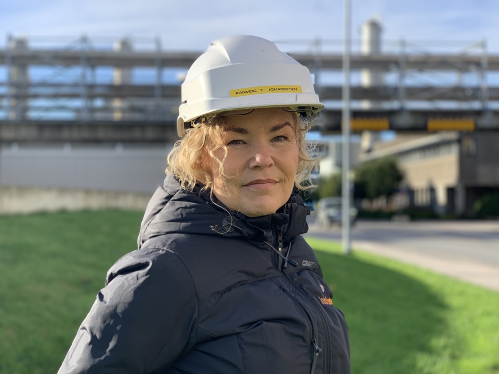 kvinne, portrett, hvit hjelm, blå jakke, industripark i bakgrunnen.