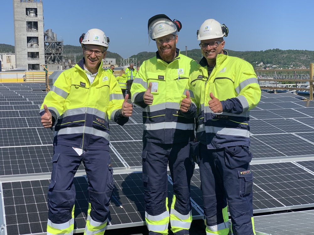 three men posing, dressed in yellow and blue working clothes, white helmets, standing on a flat roof covered with solar panels.
