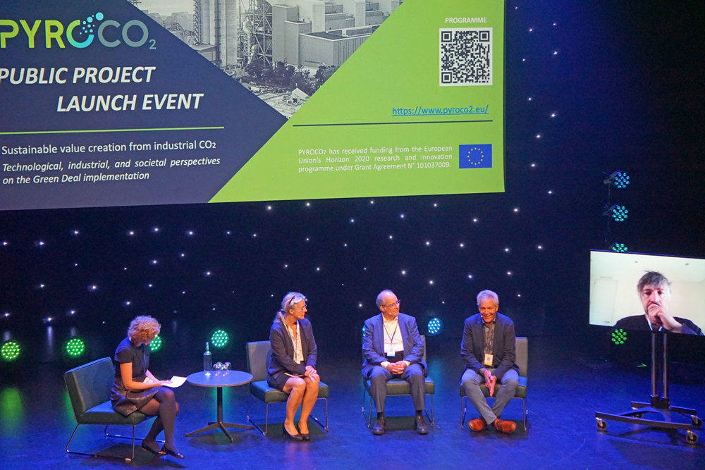 four people on stage, big presentation on screen behind them