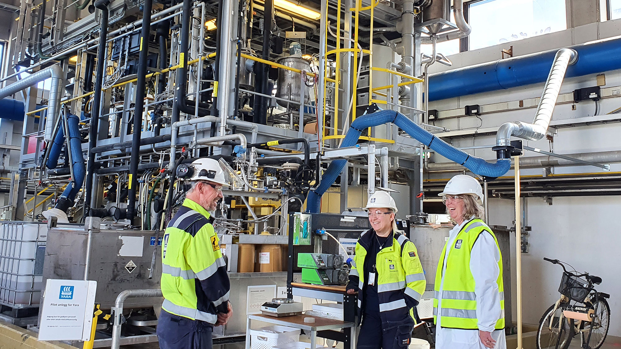 three people standing by a pilot plant, mini factory, in a big hall